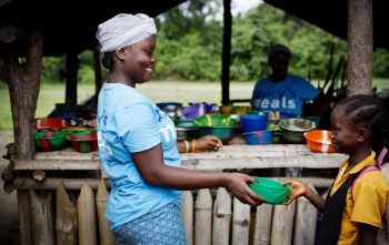 A volunteer cook is handing over a bowl of porridge to a child