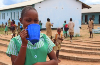 A girl in a green shirt with a blue cup covering her mouth, she is standing outside a school with children behind her