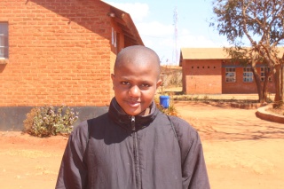 Yunus standing in a black jacket in front of his school building in Malawi