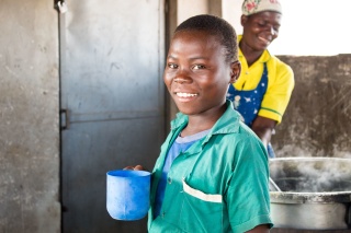 child in kitchen holding a mug, they are smiling. A woman cooks in a large stove behind the child.