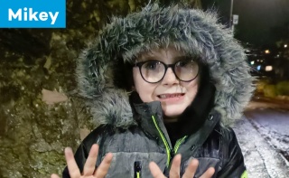 A boy stands outside at night with his hands showing a 'ten' sign. He has a fuffy hood over his hair. The text reads 'Mikey' 