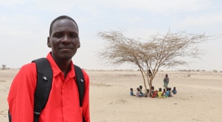 Robert, a teacher in Kenya, wearing a red shirt and backpack looking to camera with a tree and class of children in the background