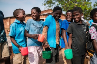 Children laughing and smiling, mugs in hand, as they queue for food in Malawi