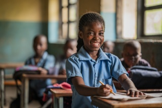 A smiling girl sits at a classroom desk pen in hand