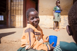 A smiling child sits outside with a mug of Mary's Meals porridge